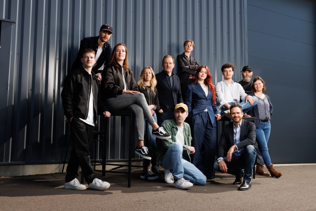 Photographie de l'équipe complète de l'agence Nesso posant devant un bâtiment industriel, intégrée dans un montage graphique avec des formes terracotta sur fond bleu clair.